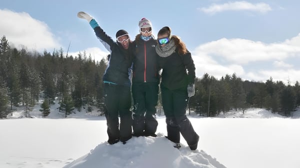 Drei Personen in warmer Winterkleidung stehen mit erhobenen Armen in einem verschneiten Wald auf dem Gelände des Appleby College.