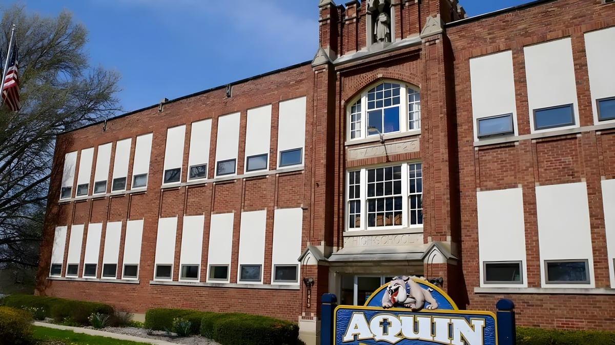 Das mehrstöckige Backsteingebäude der Aquin High School ist von Bäumen umgeben und zeigt eine amerikanische Flagge.