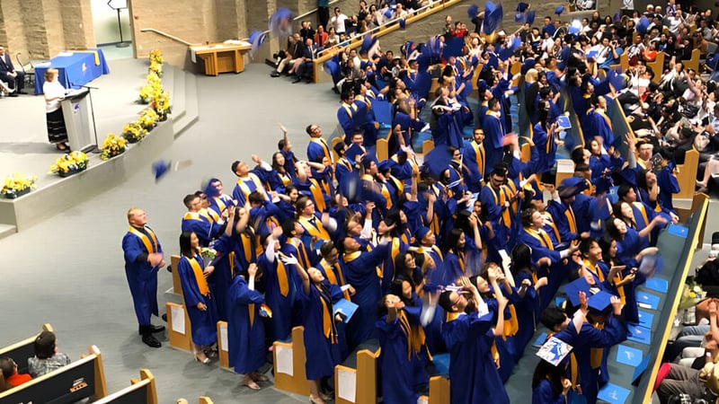 Eine große Gruppe von Absolventinnen und Absolventen in blauen und goldenen Roben versammelt sich in der Aula der A.R. MacNeill Secondary School während einer Abschlussfeier.
