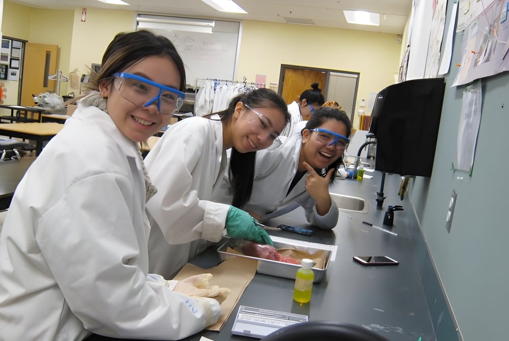 Drei Schüler der A.R. MacNeill Secondary School arbeiten in weißen Laborkitteln gemeinsam an einem Arbeitsplatz im Labor.