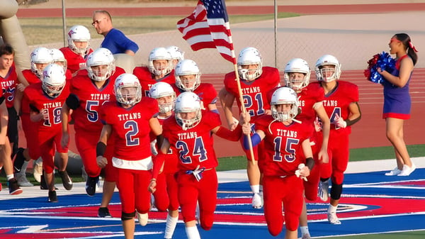 Eine Gruppe junger Footballspieler in roten Trikots trägt die amerikanische Flagge beim Betreten des Spielfelds auf dem Gelände der Arcadia High School.