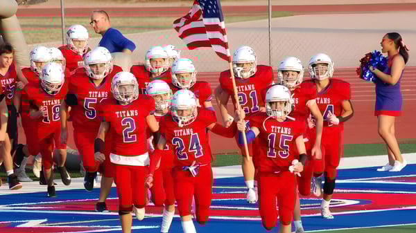 Eine Gruppe junger Footballspieler in roten Trikots trägt die amerikanische Flagge beim Betreten des Spielfelds auf dem Gelände der Arcadia High School.