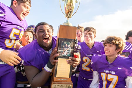 Schüler der Archbishop Riordan High School feiern mit einer Trophäe vor einem Stadion unter blauem Himmel.
