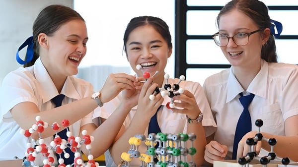 Drei Schülerinnen der Ardscoil Mhuire untersuchen lachend bunte Molekülmodelle in einem Laborraum.