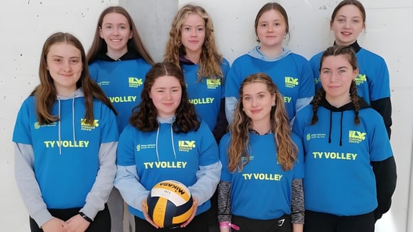 Eine Gruppe junger Volleyballspielerinnen in blauen Trikots posiert vor einer weißen Wand auf dem Campus der Ardscoil Mhuire.
