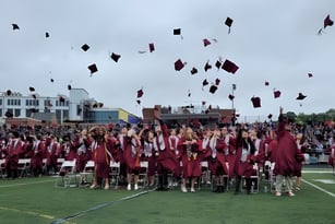 Eine große Gruppe von Absolventen der Arlington High School wirft ihre Hüte bei der Abschlussfeier auf einem Feld hoch.