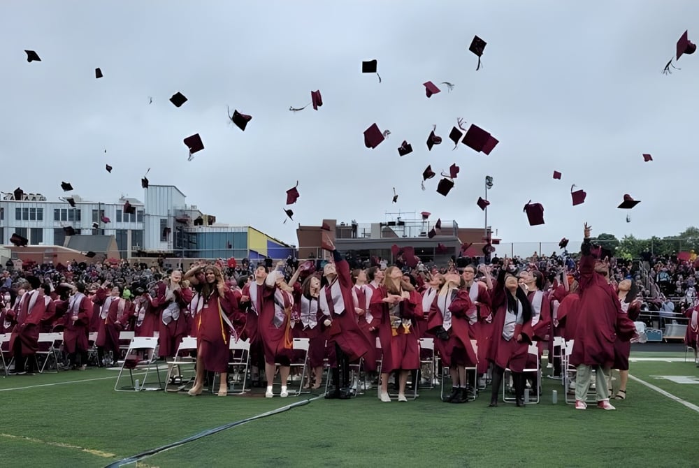 Eine große Gruppe von Absolventen der Arlington High School wirft ihre Hüte bei der Abschlussfeier auf einem Feld hoch.