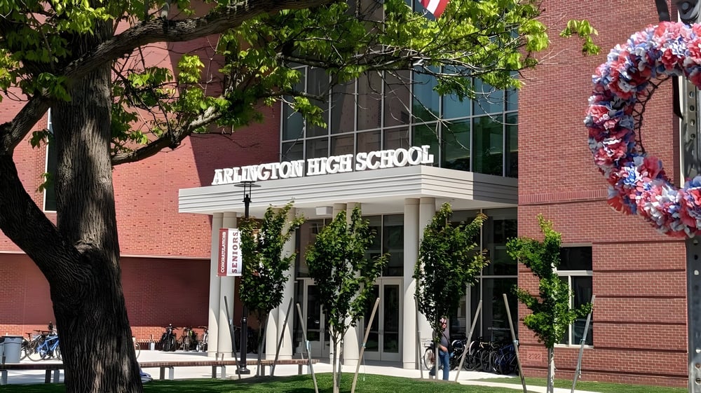Das Backsteingebäude der Arlington High School ist von grüner Vegetation umgeben und zeigt ein farbenfrohes Banner im Vordergrund.