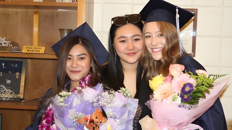 Drei Absolventinnen der Arroyo Pacific Academy in Talaren halten Blumensträuße in einem häuslichen Umfeld.