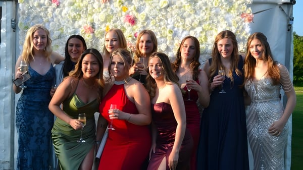 Eine Gruppe junger Frauen in Abendkleidern steht vor einer farbenfrohen Blumenwand auf dem Campus der Ashford School.