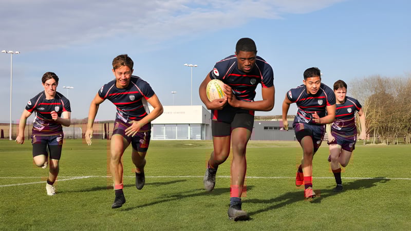 Schüler der Ashford School spielen Rugby auf einem Rasenfeld vor einem Sportgebäude.