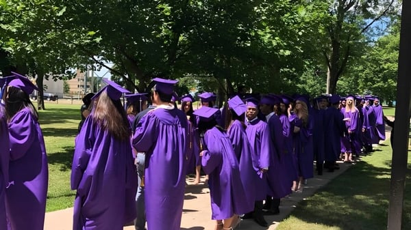 Eine Gruppe von Absolventen in lila Roben und Mortarboards geht auf dem grünen Campus der Assumption College Catholic High School.
