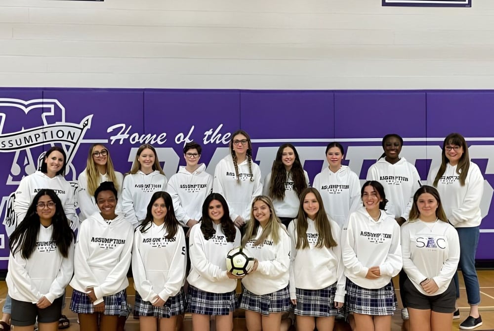 Eine Gruppe Schülerinnen in weißen Uniformen steht vor einem lila Banner auf dem Campus der Assumption College Catholic High School.