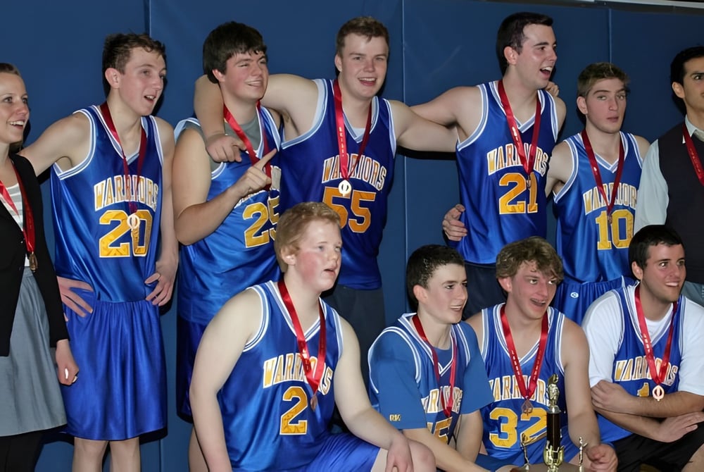 Eine Gruppe junger Basketballspieler der Athens District High School mit blauen Trikots steht vor einer blauen Wand und hält Medaillen.