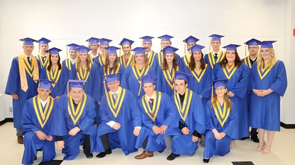 Eine Gruppe von Absolventinnen und Absolventen der Athens District High School steht in blauen und gelben Talaren vor einem weißen Hintergrund.