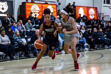 Zwei Schülerinnen des Athlete Institute spielen Basketball auf dem Spielfeld vor Zuschauern.