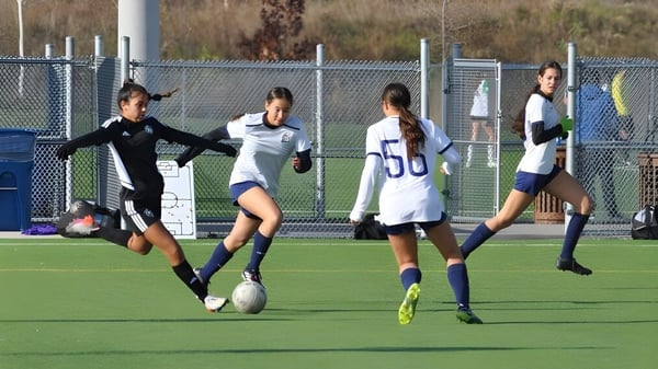 Eine Gruppe von Fußballspielerinnen trainiert auf dem Feld des Athlete Institute vor einem Zaun und Gebäuden.