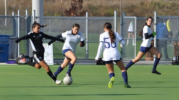 Eine Gruppe von Fußballspielerinnen trainiert auf dem Sportplatz des Athlete Institute vor einem Zaun und Gebäuden.
