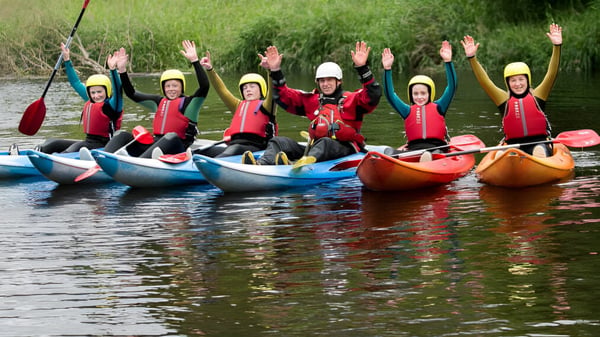 Eine Gruppe von Schülerinnen und Schülern des Athlone Community College paddelt mit bunten Schwimmwesten auf Kajaks auf einem von grüner Vegetation umgebenen Gewässer.