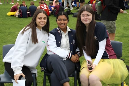 Drei Schülerinnen sitzen auf dem Grasfeld auf dem Campus des Atwell College mit Menschen im Hintergrund.
