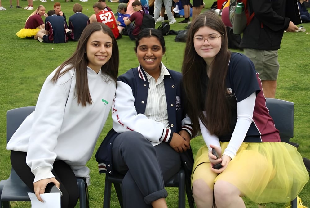 Drei Schülerinnen sitzen auf dem Grasfeld auf dem Campus des Atwell College mit Menschen im Hintergrund.