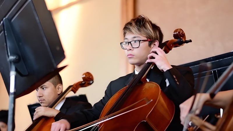 Ein Schüler der Auburn Adventist Academy spielt Cello auf der Bühne mit weiteren Musikern im Hintergrund.