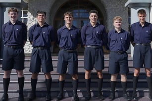 Eine Gruppe von sechs Schülerinnen und Schülern in blauen Uniformen steht vor einem steinernen Gebäude mit Rundbogenfenstern auf dem Campus der Auckland Grammar School.