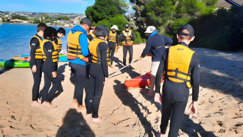Eine Gruppe von Schülern des Avenues College steht in Neoprenanzügen am Strand nahe einem Gewässer.