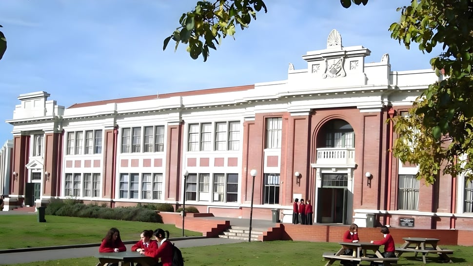 Das historische Hauptgebäude der Avonside Girls' High School steht auf einer Wiese, umgeben von Bäumen und Schülerinnen auf Bänken.