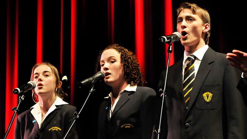 Drei Schülerinnen und Schüler von Awatapu College treten auf der Bühne vor roten Vorhängen auf und singen in Mikrofone.