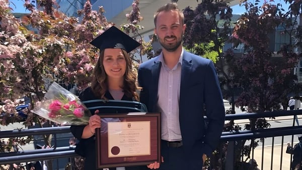 Eine Frau im Abschlusskleid und ein Mann im Anzug halten bei der Baccalieu Collegiate ein Diplom vor blühenden Bäumen.