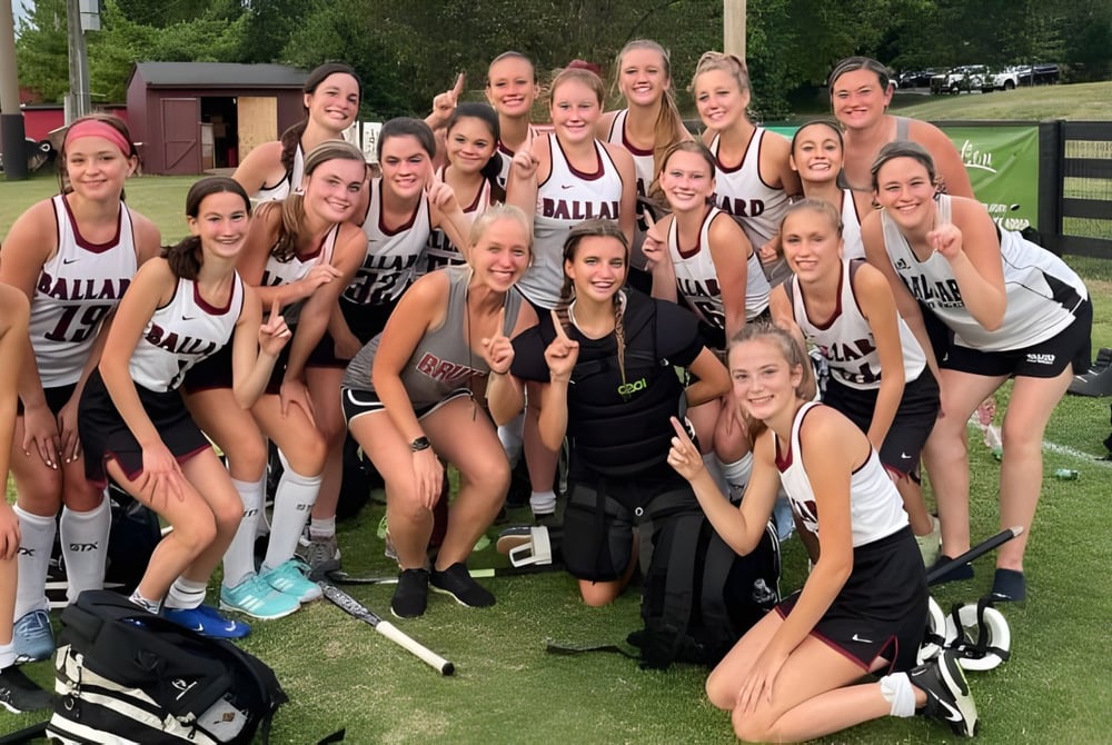 Eine Gruppe junger Athletinnen der Ballard High School steht gemeinsam auf einem Rasenfeld vor einem Gebäude und Bäumen.