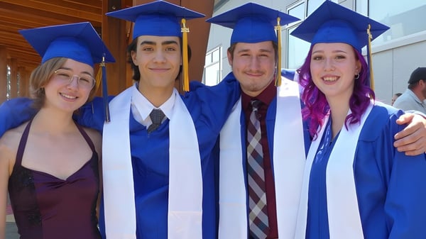 Vier Absolventen der Ballenas Secondary School posieren gemeinsam in ihren Abschlusskleidern in einer schulischen Umgebung.