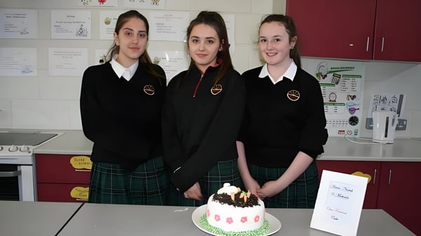 Drei Schülerinnen der Ballinteer Community School stehen gemeinsam im Klassenzimmer vor einem Geburtstagskuchen auf dem Tisch.