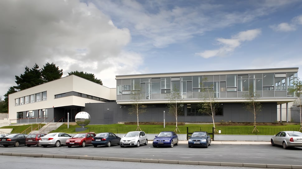 Das moderne Gebäude des Ballybay Community College mit Glasfassade und einem Parkplatz im Vordergrund.
