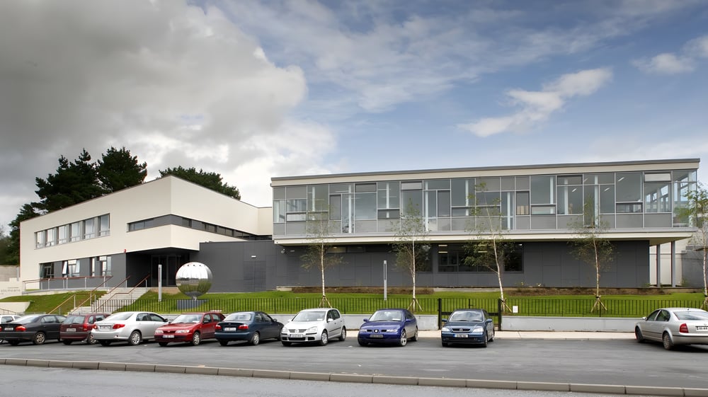 Das moderne Gebäude des Ballybay Community College mit Glasfassade und einem Parkplatz im Vordergrund.