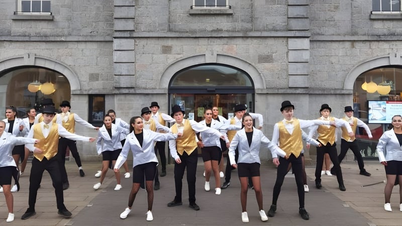 Eine Gruppe von Schülern der Ballymahon Vocational School führt vor einem Steingebäude eine synchronisierte Tanzroutine in weißen und goldenen Uniformen auf.