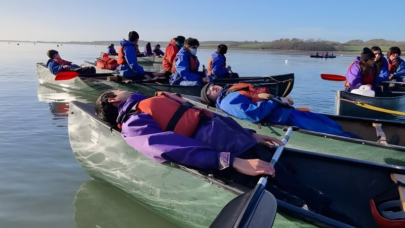 Schüler des Banagher College sitzen in bunten Rettungswesten in kleinen Booten auf dem Wasser vor einer landschaftlichen Kulisse.