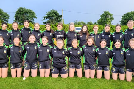 Eine Gruppe junger Fußballspielerinnen steht auf dem Sportfeld des Banagher College vor Bäumen und klarem Himmel.