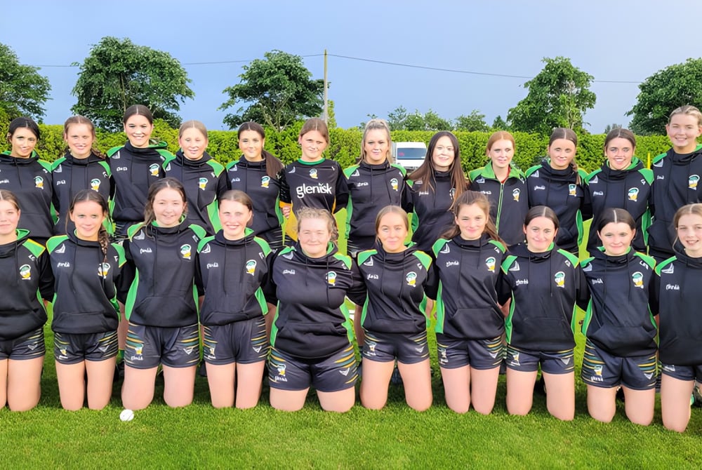 Eine Gruppe junger Fußballspielerinnen steht auf dem Sportfeld des Banagher College vor Bäumen und klarem Himmel.