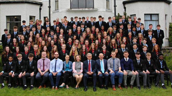 Eine große Gruppe von Schülerinnen und Schülern posiert vor dem markanten Gebäudekomplex der Bandon Grammar School für ein Gruppenfoto.