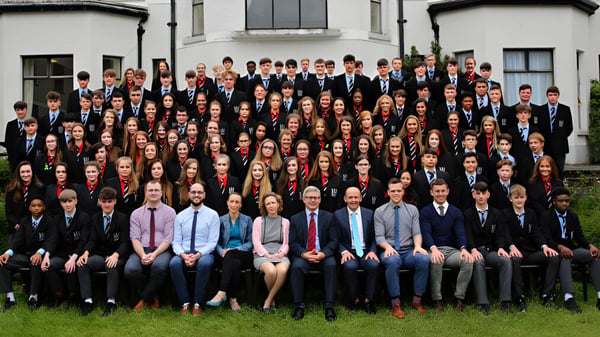 Eine große Gruppe von Schülerinnen und Schülern steht in formeller Kleidung vor einem Gebäude der Bandon Grammar School mit klassischer Architektur.