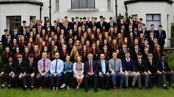 Eine große Gruppe von Schülerinnen und Schülern steht in formeller Kleidung vor einem Gebäude der Bandon Grammar School mit klassischer Architektur.