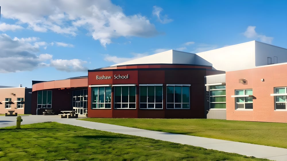 Das moderne mehrstöckige Schulgebäude der Bashaw School mit rotem Eingang steht auf einer Rasenfläche unter blauem Himmel mit weißen Wolken.