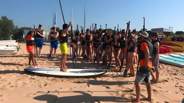 Schüler des Baxter College stehen am Strand mit Wassersportausrüstung wie Surfbrettern und Kajaks.