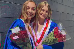 Zwei Absolventinnen in bunten Abschlussgewändern mit Blumensträußen vor einer grauen Betonwand an der Bear Creek Secondary School.
