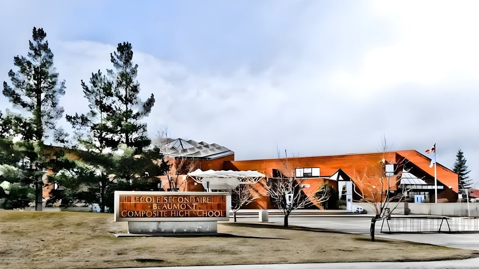 Das moderne orangefarbene Gebäude der École Secondaire Beaumont Composite High School steht umgeben von Kiefern und einer Wiese unter bewölktem Himmel.