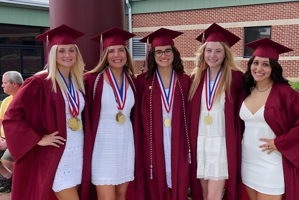 Fünf Absolventinnen in roten Talaren der Beaver Area High School stehen mit Diplomen vor einem Backsteingebäude.