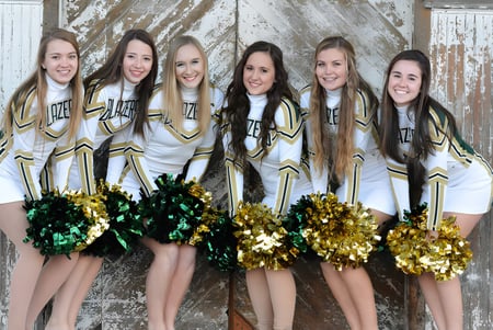 Eine Gruppe Cheerleader der Beckman Catholic School posiert mit gold-grünen Pompons vor einem verwitterten Holzhintergrund.