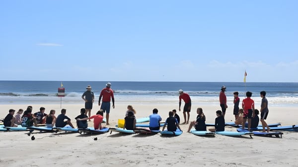 Schüler der Bedford School stehen mit ihren Surfbrettern am Strand vor dem Ozean.
